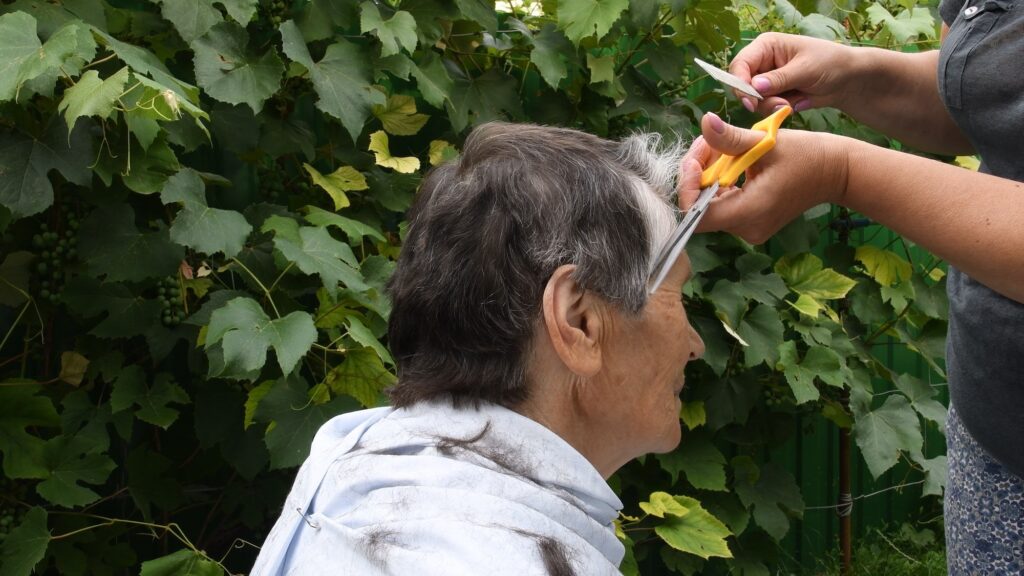 Hands trim hair on temple of old lady with half gray haired short haircut